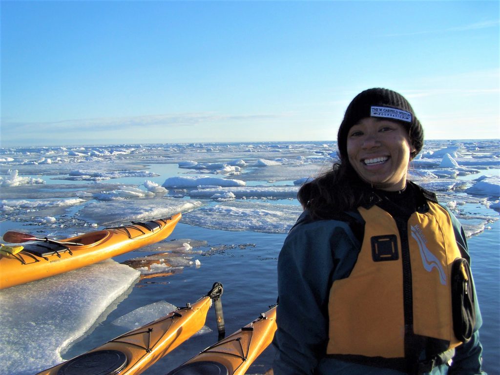 Emily Choy stands in front of artic ice and ocean wearing a yellow lifejacket, alongside three yellow kayaks
