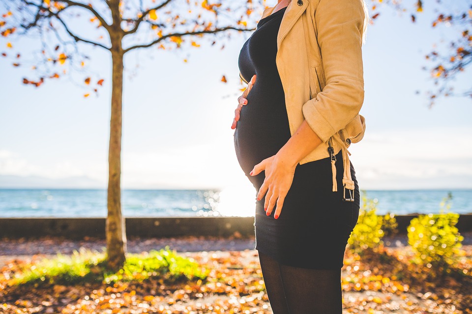 Pregnant woman standing in front of water, tree in background, during Autumn.