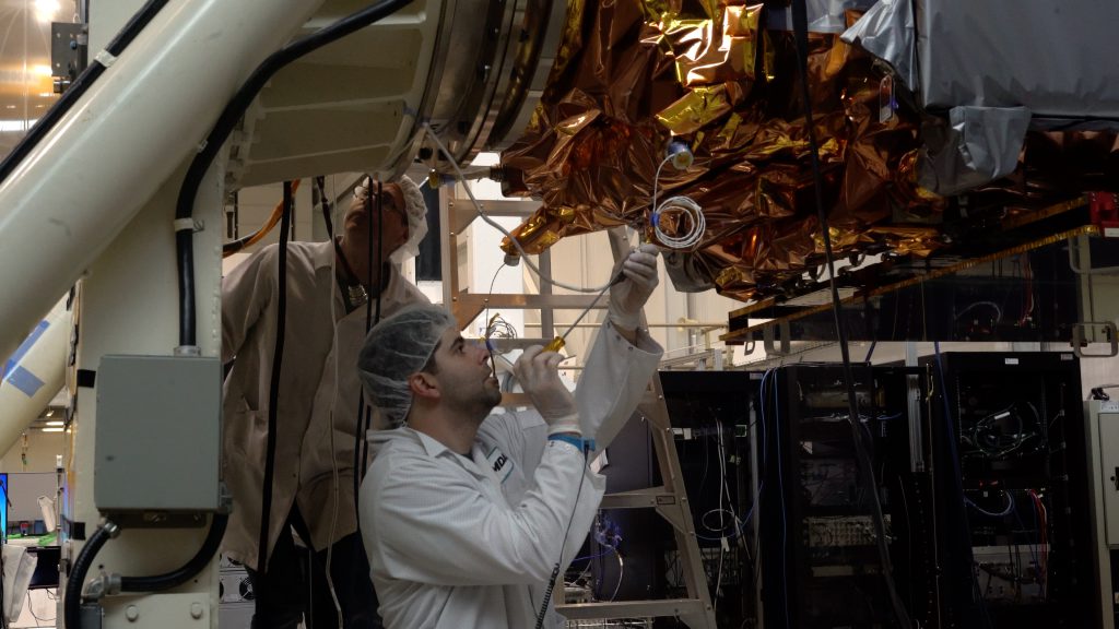 A worker in a flightsuit makes adjustments to a satellite