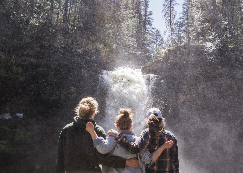 Three hikers stand with arms around each other looking at a waterfall